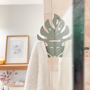 Könnte beinhalten: Ein salbeigrüner, blattförmiger Handtuchhaken hält ein flauschiges weißes Handtuch in einem Badezimmer. Der Haken ist an einer Glas-Duschabtür angebracht. Im Hintergrund befinden sich ein gerahmter botanischer Druck und Holzregale.