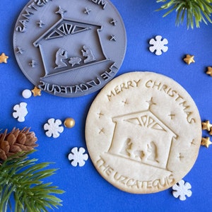 Puede incluir: Un cortador de galletas de plástico gris con las palabras "Merry Christmas The UZcatequis" y un diseño de escena de la natividad. Se muestra una galleta blanca con el mismo diseño y texto junto a ella.