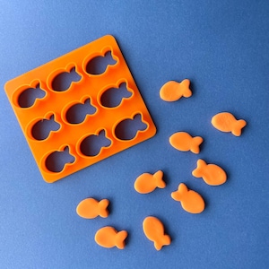 May include: Orange plastic mold with nine fish-shaped cavities, and ten orange fish-shaped candies on a blue background.