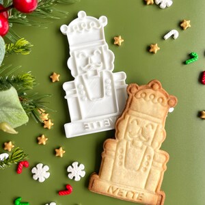 May include: A white plastic cookie cutter and a baked cookie in the shape of a nutcracker. Both have the word "Ivette" written on them. The cookie cutter and cookie are surrounded by gold stars, snowflakes, and candy canes on a green background.