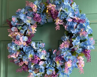 May include: A blue and purple floral wreath with a gold hanger. The wreath is made of artificial flowers and is ideal for a spring or summer door decoration.