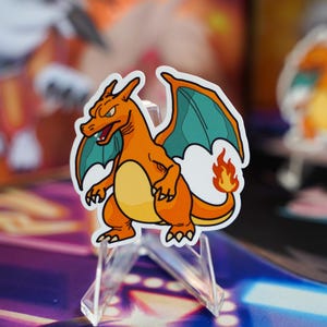 May include: A sticker of Charizard, a fiery orange and yellow dragon-like creature with teal wings and a flame on its tail. The sticker is displayed on a clear acrylic stand, with other Pokemon-themed items in the background.