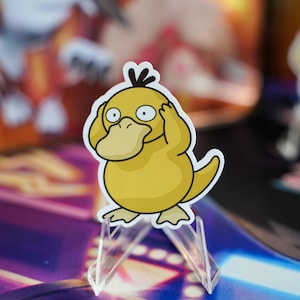 May include: A yellow Psyduck sticker with a black tuft of hair, holding its head with its hands. The sticker is on a clear acrylic stand. Other stickers are visible in the background.