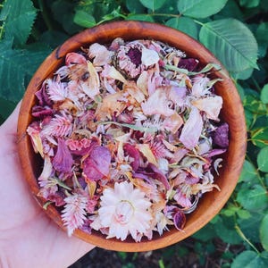 May include: A round wooden bowl filled with a mixture of dried flower petals and small blossoms in shades of pink, magenta, and cream. The collection includes various textures of dried flora, featuring a prominent light pink strawflower.