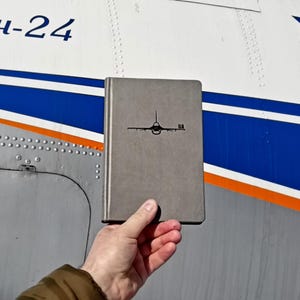 May include: A gray notebook with a black airplane silhouette and the text "UA" on the cover. The notebook is being held in front of a white, blue, and orange airplane fuselage with the text "An-24" on the side.