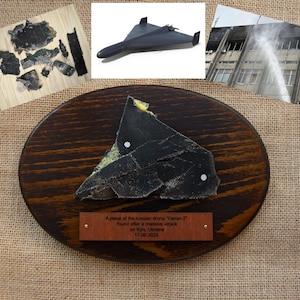 Peut inclure: Un fragment triangulaire de drone noir monté sur une plaque en bois ovale. Une plaque métallique rectangulaire porte le texte : A piece of the Russian drone Geran-2 found after a massive attack on Kyiv, Ukraine 17.09.2025. Deux photos sont incluses.