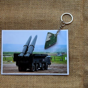 May include: A metal keychain featuring a small, triangular, green-painted fragment of debris attached to a silver-colored split ring. The keychain is displayed resting on a rectangular photograph showing a military missile launcher vehicle.