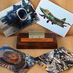 May include: A wooden display with a metal component and four images. The images show a jet engine, a military aircraft in flight, and wreckage. A wooden plaque reads "Russian SU-25 shot down in the Kherson Region, November 2022, Ukraine."