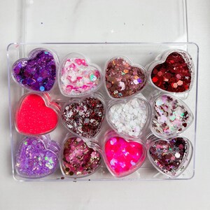 May include: A set of 10 heart-shaped containers filled with colorful glitter. The containers are arranged in two rows of five, and each container has a different color of glitter.