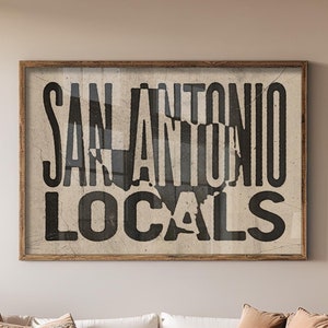 May include: A white sofa with brown and patterned pillows in a living room setting. A wooden coffee table sits in front of the sofa. A framed print with the text "San Antonio Locals" hangs on the wall behind the sofa. Two potted cactus plants are in the room.