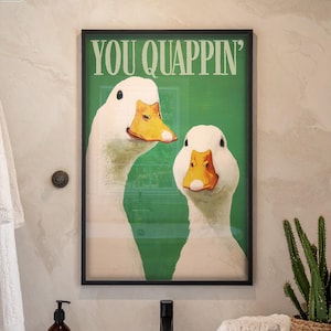 May include: A framed art print featuring two white ducks with orange beaks against a green background. The text "YOU QUAPPIN'" is above the ducks. The image is in a bathroom setting with a tub, towels, and a potted plant.