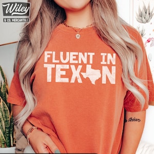 May include: A burnt orange t-shirt with the text "FLUENT IN TEXN" in white with an outline of the state of Texas. The shirt is a Comfort Colors 1717 T-Shirt in the color YAM.