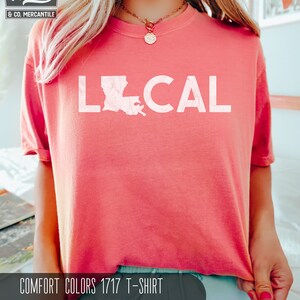 May include: A pink t-shirt with the word "LOCAL" printed on it in white. The word "LOCAL" is accompanied by an outline of the state of Louisiana. The text "COMFORT COLORS 1717 T-SHIRT WATERMELON" is printed below the graphic.