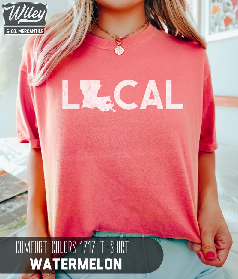 May include: A pink t-shirt with the word "LOCAL" printed on it in white. The word "LOCAL" is accompanied by an outline of the state of Louisiana. The text "COMFORT COLORS 1717 T-SHIRT WATERMELON" is printed below the graphic.