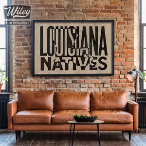 May include: A black framed print with the text "Louisiana Natives" in white letters on a beige background. The print is hanging on a brick wall above a brown leather couch.