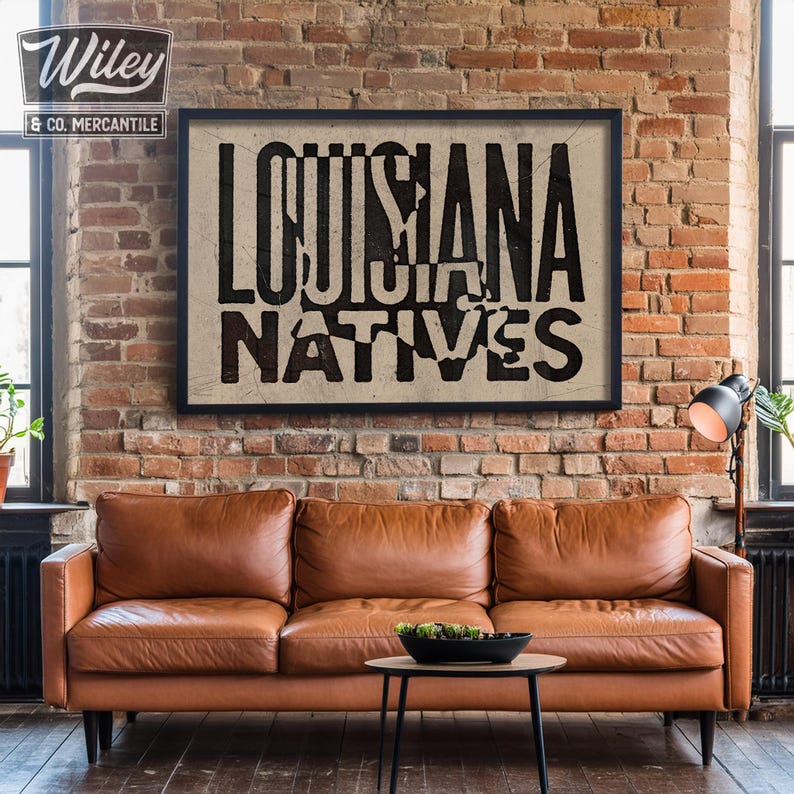 May include: A black framed print with the text "Louisiana Natives" in white letters on a beige background. The print is hanging on a brick wall above a brown leather couch.