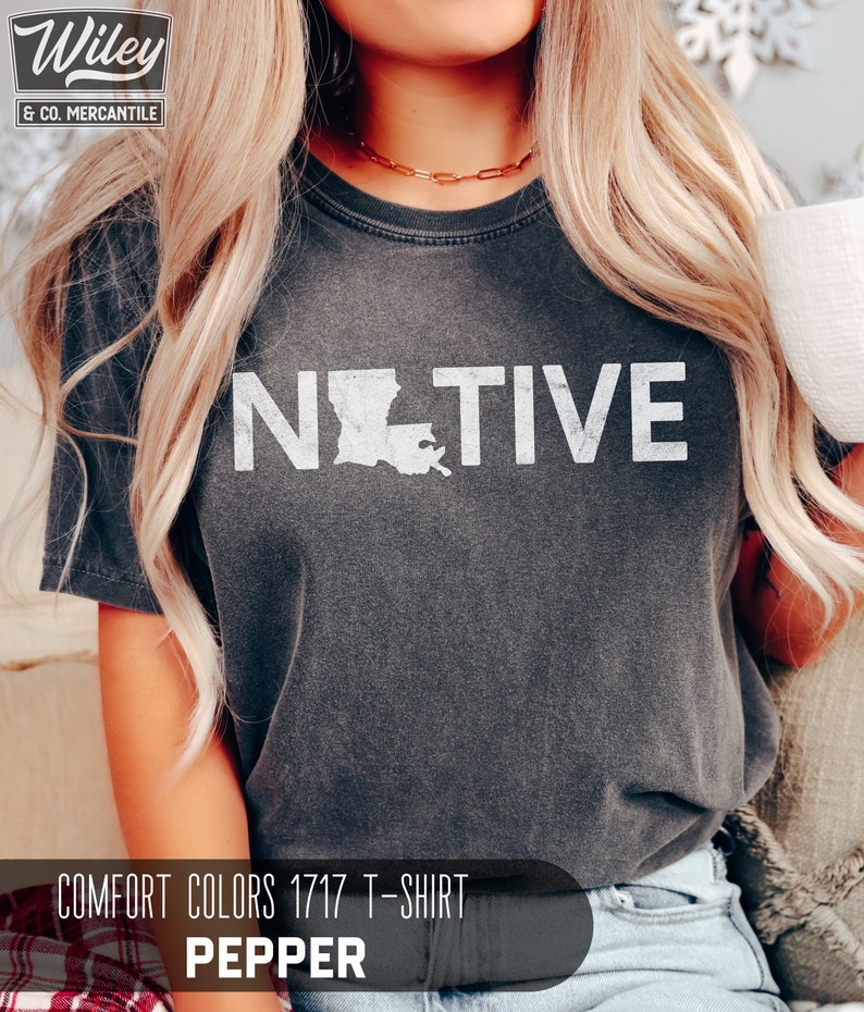 May include: Dark gray Comfort Colors 1717 t-shirt with the word "NATIVE" in white block letters, featuring the outline of Louisiana. The shirt is pepper-colored and has a vintage look. The Wiley & Co. Mercantile logo is in the upper left corner.