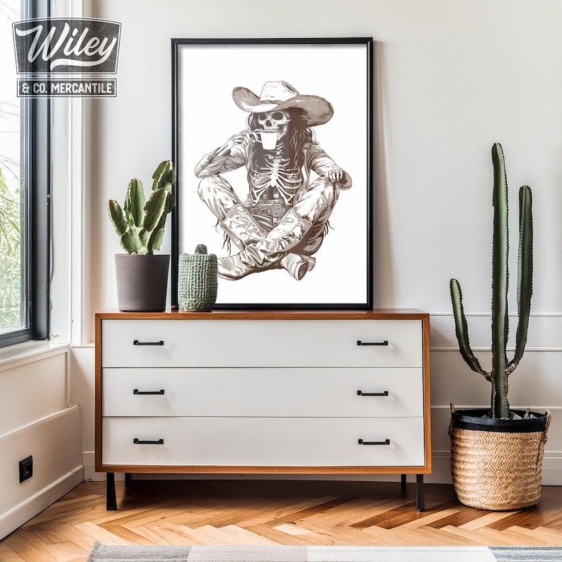 May include: A black and white illustration of a skeleton wearing a cowboy hat and drinking from a cup. The skeleton is sitting with its legs crossed and is wearing a belt with a buckle. The illustration is framed and hanging on a white wall.