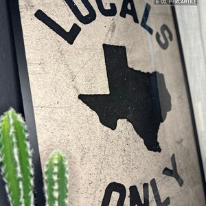 May include: A framed sign with a distressed white background and black lettering. The sign reads "Locals Only" with an outline of the state of Texas in the center. The sign is framed in black and has a small label in the top right corner that reads "Wiley & Co. Mercantile".