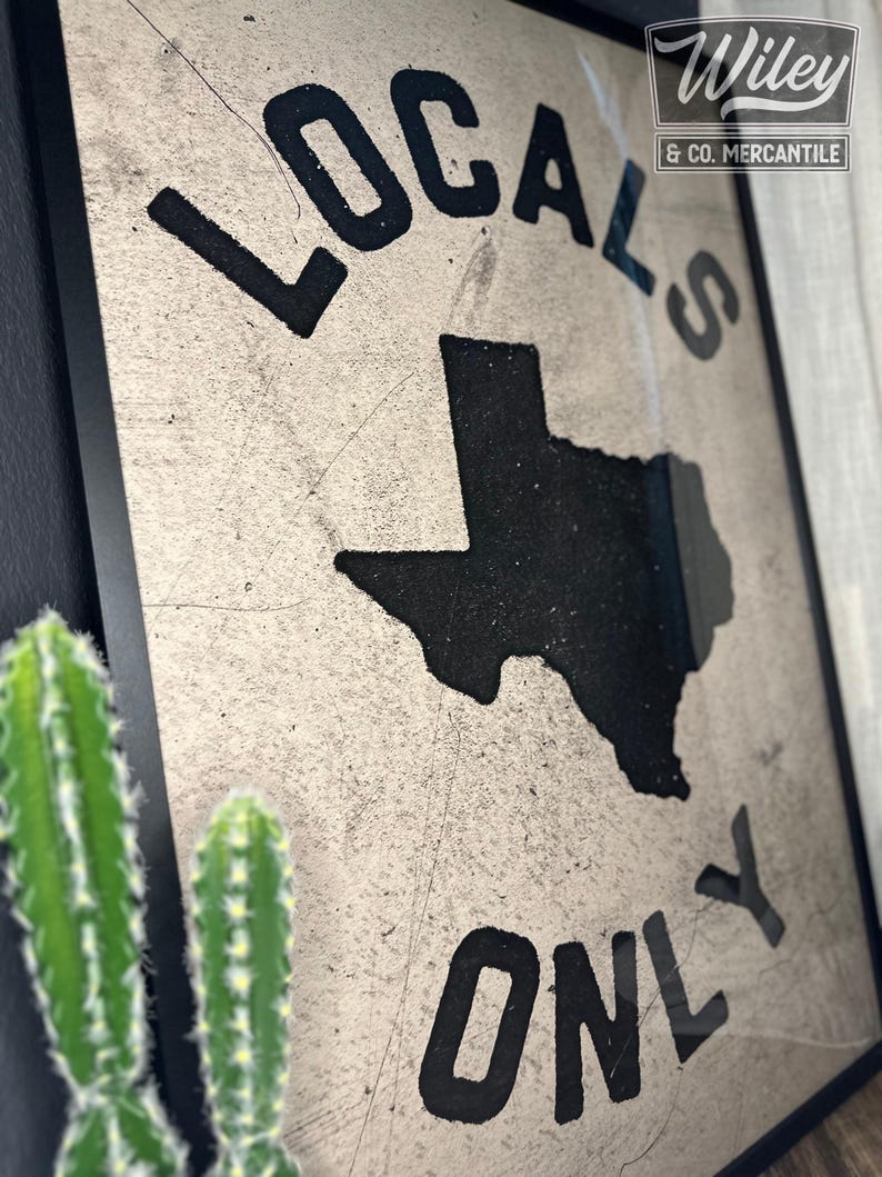May include: A framed sign with a distressed white background and black lettering. The sign reads "Locals Only" with an outline of the state of Texas in the center. The sign is framed in black and has a small label in the top right corner that reads "Wiley & Co. Mercantile".