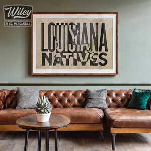 May include: A framed print with the text "Louisiana Natives" in black lettering on a beige background. The print is hanging on a sage green wall above a brown leather sectional sofa with pillows.