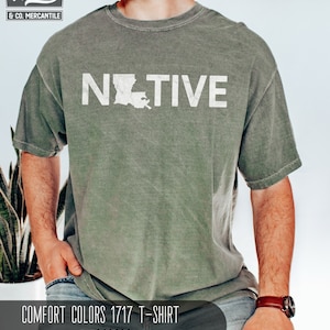 May include: A moss green Comfort Colors 1717 T-shirt with the word "NATIVE" in white block letters and a white outline of the state of Louisiana. The shirt has a vintage, worn look.