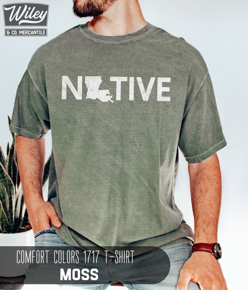 May include: A moss green Comfort Colors 1717 T-shirt with the word "NATIVE" in white block letters and a white outline of the state of Louisiana. The shirt has a vintage, worn look.