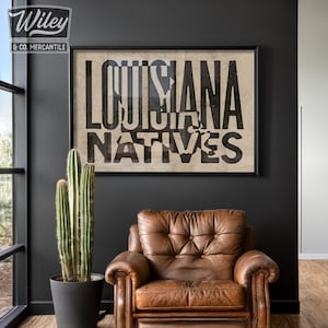May include: A black and white poster with the text "Louisiana Natives" in a distressed font. The poster is framed in black and hanging on a dark gray wall. A brown leather armchair sits in front of the poster.