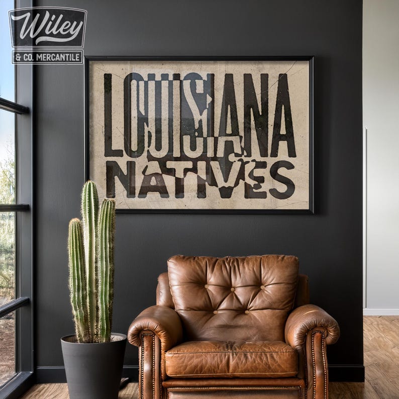 May include: A black and white poster with the text "Louisiana Natives" in a distressed font. The poster is framed in black and hanging on a dark gray wall. A brown leather armchair sits in front of the poster.
