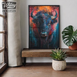May include: A framed art print featuring a colorful, abstract painting of a bison. The artwork uses shades of blue, red, and orange. The print is displayed on a wooden shelf, with a small potted plant in front of it. The frame is black.