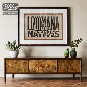 May include: A wooden cabinet with three drawers featuring a carved design. The cabinet is brown with a gold finish. A framed print with the text "Louisiana Natives" is hanging on the wall above the cabinet.