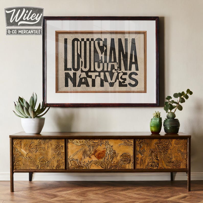 May include: A wooden cabinet with three drawers featuring a carved design. The cabinet is brown with a gold finish. A framed print with the text "Louisiana Natives" is hanging on the wall above the cabinet.