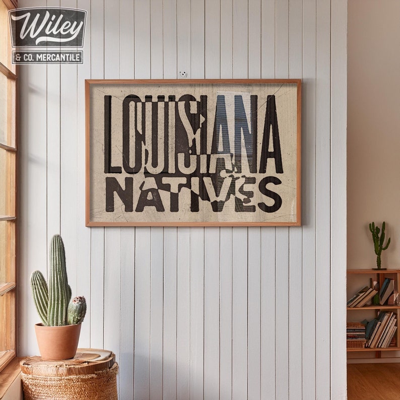 May include: A framed print with the text "Louisiana Natives" in black lettering on a white background. The print is in a wooden frame with a light brown finish.