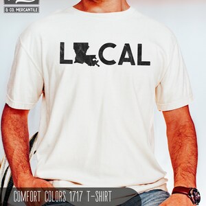May include: A white t-shirt with the word "LOCAL" printed in black with a distressed outline. The letter "O" is replaced with the outline of the state of Louisiana. The text "COMFORT COLORS 1717 T-SHIRT IVORY" is printed below the graphic.