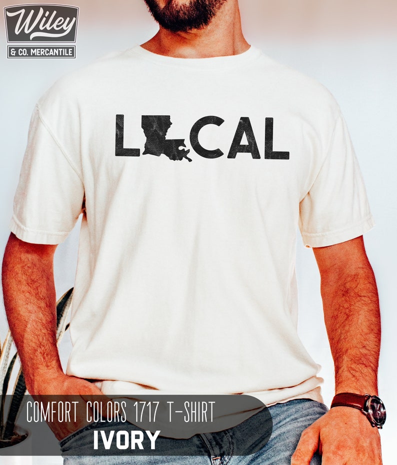 May include: A white t-shirt with the word "LOCAL" printed in black with a distressed outline. The letter "O" is replaced with the outline of the state of Louisiana. The text "COMFORT COLORS 1717 T-SHIRT IVORY" is printed below the graphic.