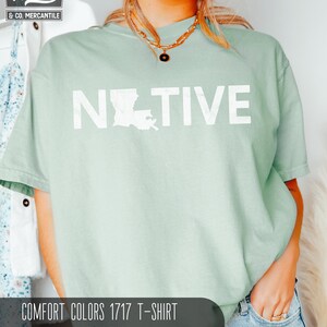 May include: A light green Comfort Colors 1717 T-shirt with the word "NATIVE" printed in white, with the outline of Louisiana replacing the "O". The shirt is a bay color. The Wiley & Co. Mercantile logo is in the upper left corner.