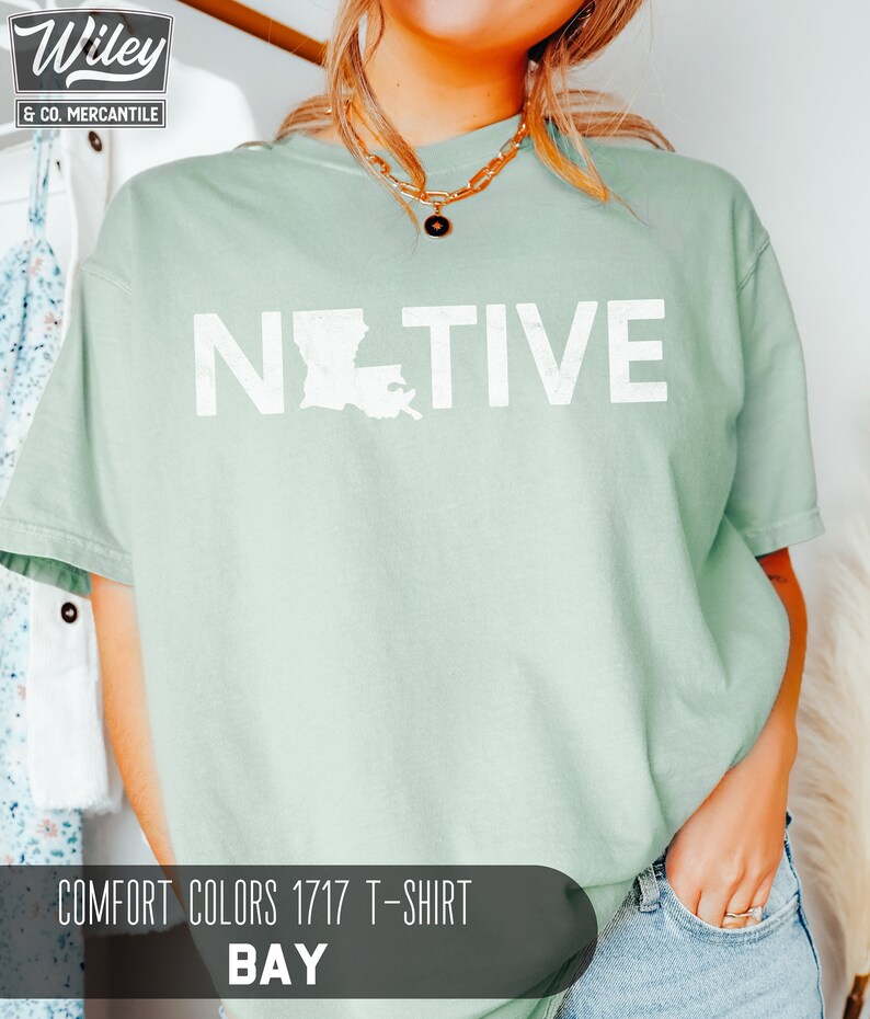 May include: A light green Comfort Colors 1717 T-shirt with the word "NATIVE" printed in white, with the outline of Louisiana replacing the "O". The shirt is a bay color. The Wiley & Co. Mercantile logo is in the upper left corner.