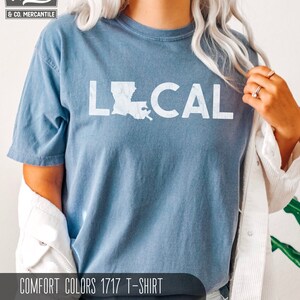 May include: A blue jean t-shirt with the word "LOCAL" printed in white with an outline of the state of Louisiana. The shirt is a Comfort Colors 1717 t-shirt.