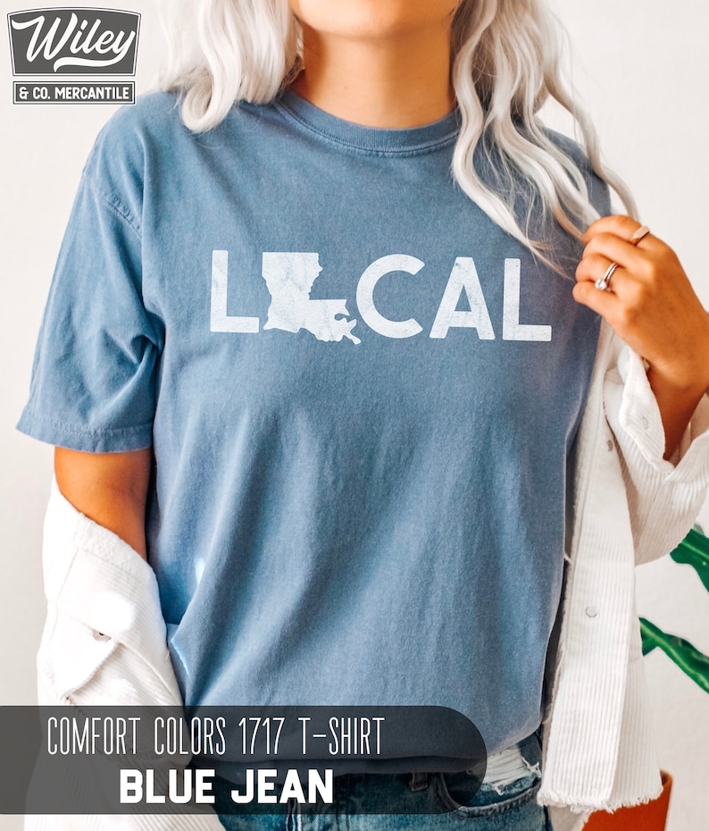 May include: A blue jean t-shirt with the word "LOCAL" printed in white with an outline of the state of Louisiana. The shirt is a Comfort Colors 1717 t-shirt.