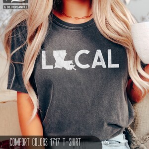 May include: A dark gray t-shirt with the word "LOCAL" printed in white, featuring an outline of the state of Louisiana. The shirt is labeled "COMFORT COLORS 1717 T-SHIRT PEPPER".