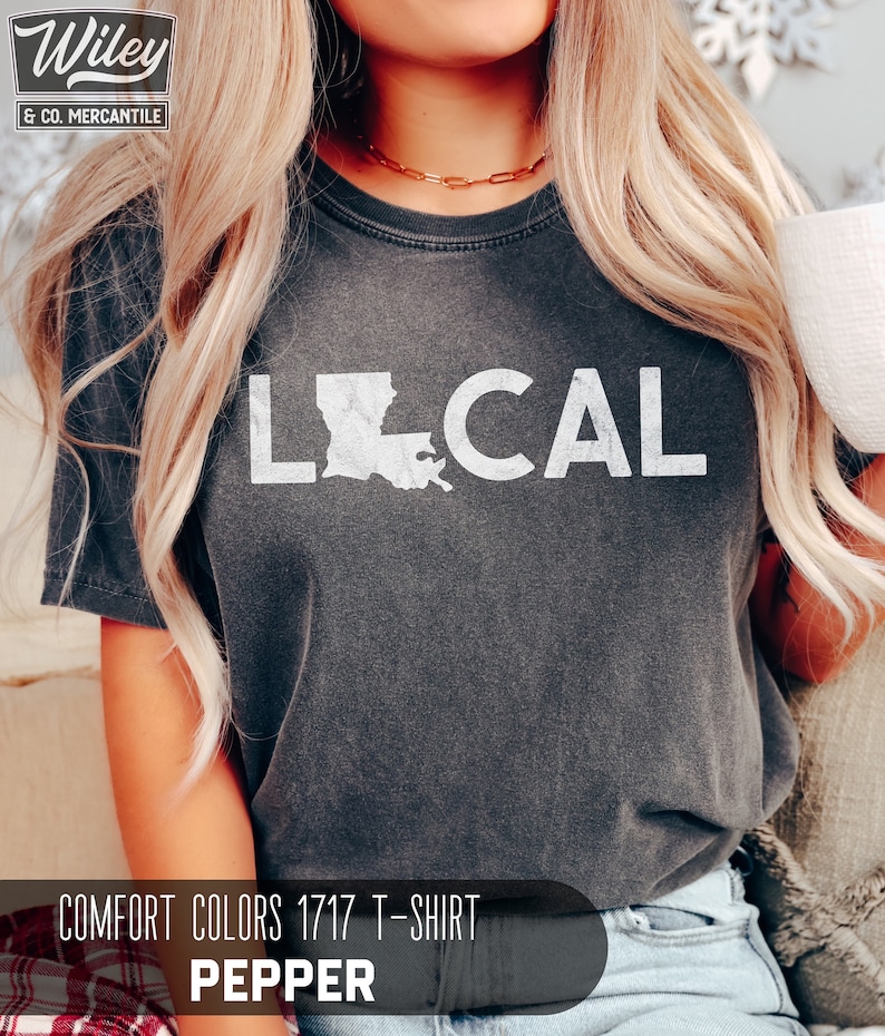 May include: A dark gray t-shirt with the word "LOCAL" printed in white, featuring an outline of the state of Louisiana. The shirt is labeled "COMFORT COLORS 1717 T-SHIRT PEPPER".