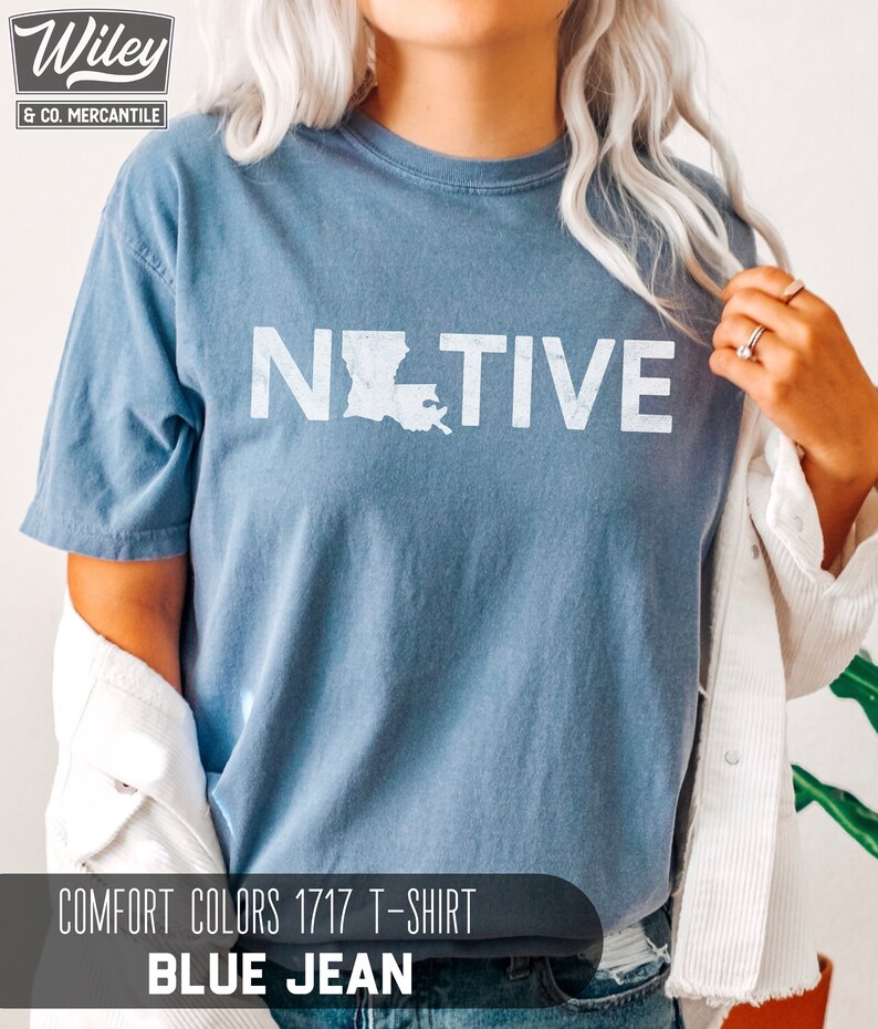 May include: Blue Comfort Colors 1717 T-shirt with the word "NATIVE" in white block letters and a white outline of the state of Louisiana. The shirt is a blue jean color and has short sleeves.