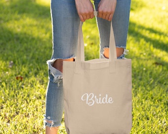 May include: A beige tote bag with the word "Bride" in white script. The bag has two handles and is held by a person wearing blue jeans. The background is a green grassy area with sunlight.
