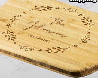 May include: A personalized bamboo cutting board with the text "The Johnsons Established 2022" surrounded by a decorative border of leaves and stars.