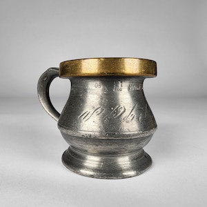 May include: A small, pewter-colored mug with a gold-colored rim and handle. The mug has a rounded body and a flared base. The mug has the number "09" and the word "HALF" etched into the side. The mug is likely an antique or vintage item.