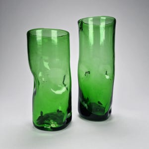 May include: Two handcrafted, green glass tumblers. The glasses have a unique, organic shape with a slightly irregular form. The vibrant green color and the imperfect shape give them a rustic, artisanal feel. The glasses are suitable for serving beverages.
