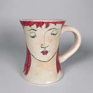May include: Cream-colored ceramic mug featuring a stylized face design. The face has closed green eyes, red hair, and red lips. A small gold-colored dot is on the nose. The mug has a curved handle and a flared rim.