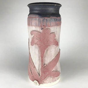 以下が含まれることがあります： 円筒形のハンドメイドセラミック花瓶。 白とピンクの背景に、ピンクのユリの浮き彫りデザインが施されています。 リムは濃い青色です。 花瓶の高さは約25cmです。