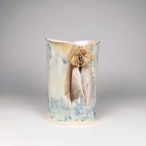 May include: A ceramic vase with a light blue and yellow glaze. The vase has a unique shape with a slight curve in the middle. It is decorated with three white feathers and a small, brown, woven piece at the top.
