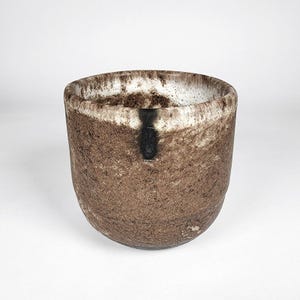 May include: A handcrafted ceramic bowl with a textured, earthy brown exterior and a white interior rim. A vertical black accent stripe is visible on the front. The bowl has a rounded base and a wide opening, suitable for serving or display.